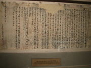  A tattered, ripped and crumpled piece of light coloured paper with writings penned in Chinese chearacters in dark ink, pinned on a wall for display in a museum.