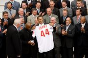 Several men of varying ages wearing suits clap as a man in the center of the photo smiles holding up a white baseball jersey which reads "OBAMA 44" on the back.