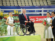 A man seated in a wheelchair on a podium, to his right and left are women.  There is also a man on the extreme right of the image.  The woman on the right holds a silver tray with a bouquet of flowers.