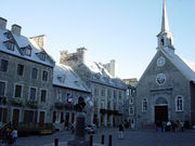 A cobblestone square with a statue, surrounded by old-fashioned stone buildings. A group of people are being photographed in front of one of the buildings