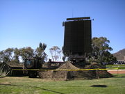 A green truck with a large rectangular object mounted on its tray. The truck is covered in camouflage netting and is surrounded by a low fence.