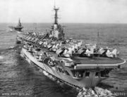 Photograph of an aircraft carrier from behind. Numerous aircraft with their wings folded are sitting on the flight deck. A second aircraft carrier is in the background, leading the first.