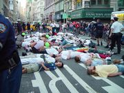 Street protest scene; people deliberately lying down on a busy city street, surrounded by onlookers and police