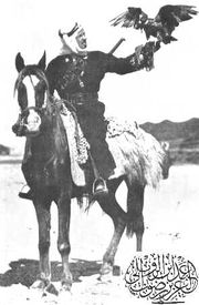 A black and white photograph of a mounted man on a dark horse. A hawk is perched on the man's outstretched hand.