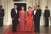 Ronald Reagan, Margaret Thatcher, Nancy Reagan and Denis Thatcher smile while standing on a red carpet leading to a large door. Men in US military dress stand in the background. Beside the door is a Union Jack flag.