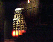 In a dark basement, a white Dalek (see previous description) appears to levitate up a small staircase of approximately seven stairs. The body of the Dalek is white, with shiny gold vertical slats and gold balls on its lower half. There is an orange-yellow glow at the Dalek's base.