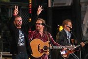 Starr is on stage with two guitarists and two microphones. He is wearing sunglasses and a black and silver t-shirt, and both of his arms are raised. His right arm forms a V-shaped peace symbol.