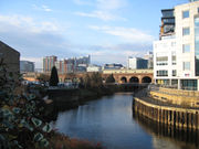 The canalised River Aire flows from the Dark Arches under Leeds's main railway station towards the bottom of the picture. To the left of the river is the lock which links the river with the Leeds and Liverpool Canal. To the right is a riverside walk beneath modern buildings, and in the distance, beyond the railway viaduct and station, are more high-rise modern buildings located on the west side of the city centre.