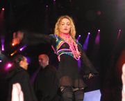 Picture of a middle-aged blond woman wearing a long-sleeve translucent black dress over black shorts and a top, the dress reaching down to mid-thigh. Brightly colored scarves are tied around her middle and shoulders. Around her neck she wears a number of colorful strings as necklaces. She stands with her hands stretched sidewards. Behind her, two men are seen facing each other.