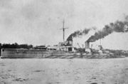 A large gray ship slowly sails through calm waters close to shore. The two funnels in the center of the ship emit clouds of dense smoke.