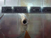 An image of a flat metallic structure with a half-inch hole punched through it. A ruler is visible to demonstrate scale.