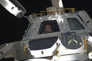 A small, squat module with three of seven windows visible, seen against the backdrop of space. Open shutters are visible next to each window, and an astronaut can be seen inside the module through the windows.