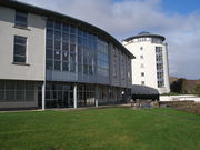 A modern 3 story building with a prominent frontage of numerous windows and constructed from a white material curves gently away from a green lawn in the foreground. In the background there is a tall white tower of a similar construction.