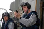 Two people wearing grey uniforms, blue life preservers and black helmets standing against a grey background. The man closest the camera is holding a pistol.