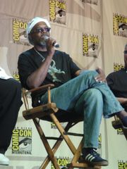 A man is sitting in a director's chair on a stage with two partially cropped out people sitting in the same type of chair on his left and right. The man is speaking into a microphone he is holding and is wearing tennis shoes, blue jeans, a black t-shirt with the film's poster image on it, sunglasses, and a white hat. In the background is a patterned design with the logo for Comic-Con.