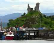 The ruins of an old building sit on top of a prominent hillock that overlooks a pier attended by fishing boats.