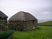 A small stone building with a thatched roof sits in a field overlooking the sea. Several large rusty metal circular rims sit against one wall and there is a partial view of another, similar building and a green rowing boat to the left.