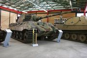 A three quarters view of a large tank with a flat-faced turret, dull yellow, green and brown wavy camouflage, on display inside a museum. The frontal armor is sloped. The long gun overhangs the bow by several meters. Two waist-high cartridges sit on their bases infront of it.