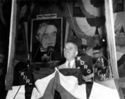 Man in wire glasses making speech behind flag-draped podium and radio microphones