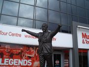 Statue of a man with a scarf around his neck and his arms aloft. Behind the statue is the exterior of a stand.