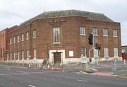 A plain corner building of the 1930s on a main road. Traffic-lights, railings, bollards and pedestrian crossings with tactile paving can also be seen. The frontage of the building is mainly brick, but the lowest course, of Portland stone, extends as far as the sills of the eleven ground-floor windows (eight on the main road and three on the side road to the right). The wooden double entrance door set diagonally on the corner also has a stone surround. Above it is a long window with a projecting brick surround, and above this is a large sign in capital letters, originally reading SHEEPSCAR but now with the second "E" missing. The upper floor has smaller windows above all of the ground-floor ones except for the furthest one on the main road, where there is no upper floor. The building has a slate half-hipped roof.