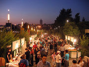 An outdoor book fair at dusk. Vendors are in booths along both sides of a walkway crowded with people.