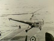 The forward section of an aircraft carrier's flight deck. A helicopter with United States markings and the letters "UP-28" painted on the side is hovering just above.