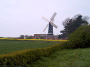 Beyond a foreground of green fields and low hedges is a tall, black cylindrical building with four white sails attached, set in a cruciform arrangement.