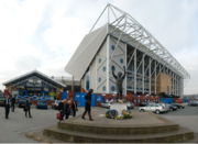 Outside a large stadium with a cantilevered stand at the right-hand end and lower buildings on the left.  Other low-rise buildings, including some houses at the front of the picture, can be seen in the foreground and background.  In the distance are some low hills with fields and some housing.