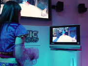 Inside a relatively dark room, a black-haired Caucasian woman turned away from the camera plays a video game which is displayed on two bright screens.