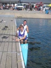 Photo of narrow kayak next to wooden dock. Two people sit in the kayak, each holding a paddle.