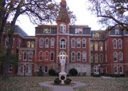 A statue sits in front of a large four-story red brick building. To the right of the entrance, the building goes in a little ways, and then back out. To the left of the entrance is a small two-story area connecting the main part of the building to the rest of it. Above and behind the two-story part is a four-story part of the building