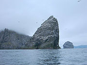 A large, triangular rock rises from the misty waters, with more islands behind and Northern Gannets flying around it.