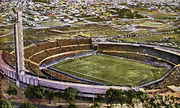 Painting of a bowl-shaped stadium, from a viewpoint high above. The stadium contains a football pitch. There are figures on the pitch, but they are too small to make out any detail. The stands, open to the air, are all full. At the left of the stadium there is a large white tower. In the foreground in front of the stadium is a row of trees. Beyond the stadium in the background is an indistinct mix of fields and buildings.