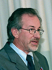 A man with dark grey hair and a light grey beard wearing a pair of glasses, grey suit, and dark striped tie.