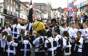 A street lined with shops is filled with hundreds of people. In the foreground are children wearing black vests each one defaced with a large white cross. The children surround a fiddler. In the background are spectators.