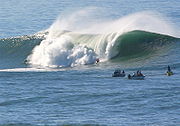 Photo of taller-than-human-sized wave breaking with several watching surfers in foreground