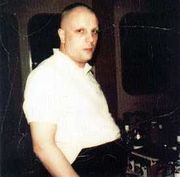 An overweight white male with shaved head and eyebrows, wearing a white short-sleeved shirt and black trousers, looks at the camera with a neutral expression.  The room behind him appears dark, and several unidentifiable pieces of equipment are visible.