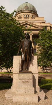 A large domed building overlooks a full-length statue of balding white male with a mustache and long goatee and wearing a knee-length coat.  The pedestal is engraved "Lawrence Sullivan Ross".