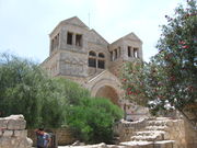 A light colored stone church with a triangular roof, with two towers flanking the entrance, in the background. In the foreground are low stone walls, a tree and some bushes