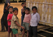 Six young children standing in front of a building with a woven wall, with a man whose head is not visible standing in the background