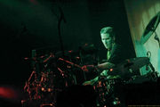 Drummer with crewcut, wearing black t-shirt, intently plays his large drum set in the spotlight.