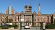 A long mid-Victorian Jacobethan-style building of red brick can be seen beyond a wide entrance with open metal gates and adjacent railings.  A car is about to go through the entry barrier.  The building has three storeys with, in the centre, a large round-headed window occupying the two storeys over the entrance and a tower above with turrets at the corners.  On either side of the tower, bay windows project forward on all three floors, and parapets and six shaped gables can be seen along the roof-line.  Behind and to the left can be seen a tall modern building, while behind and to the right there is a tall chimney.