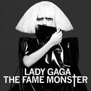 A woman in a blonde blunt bob cut. She wears a shiny black leather dress covering her body completely. With her right hand she holds the collar of the dress and uses it to cover her mouth. In front of her, the words "Lady Gaga" and "The Fame Monster" are written in white capital font, with the T of Monster being stylized as a cross (†).