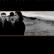 A wide monochrome photograph of four men standing in the desert sits slightly above the center of black image, spanning the width of the background. The men are standing on the left half of the photograph, while a mountain range is visible on the right half. Small gold stripes border the top and bottom of the photograph. Tiny gold text reading "THE JOSHUA TREE U2" is stretched across the top of the cover against the black background.