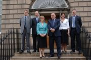 Four men and two women in smart dress pose on steps to large town building.