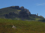 A grassy moorland in the foreground with a complex of  high, grey cliffs at centre under a blue sky. A tall rock pinnacle stands under the cliffs at right.