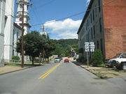 A highway runs toward a bridge between an ornate building with a clock tower and a plain three-story red brick building. A forested hill rises on the far side of the bridge, which is decorated with U.S. flags on poles. A double highway sign along the road indicates that Highway 120 turns to the left (west) before the bridge and that Highway 664 continues across the bridge and heads generally north. Several cars are moving along the highway and over the bridge.