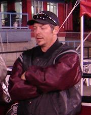 Half-length view of a person in his early 40s.  He is standing upright with his arms folded across his chest.  He is wearing a black and maroon coat and a baseball cap.