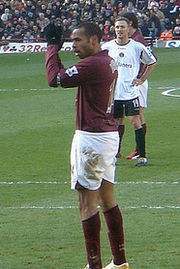 A man in association football attire comprising dark red shirt and white shorts claps his hands. Two other players, one in a white shirt and dark shorts and one in a dark red shirt and white shorts, are visible in the background, as is a crowd of spectators.